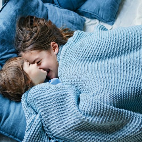 Happy siblings snuggling under warm knitted blanket. Little boy and his teenager sister enjoying staying at home at cold winter day having fun together, Family quality time on holidays.